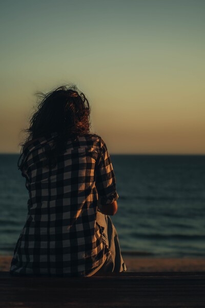 Show back of woman with long brown hair sitting on beach looking out over lake towards horizon.